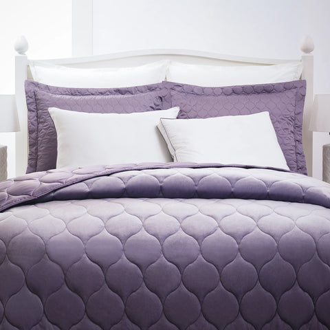 Two lavender pillows large, stand tall on the white headboard of a bed. With two right pillows in front, the left pillow is straight, while the right pillow leans on the left. In front of the pillow is a lavender comforter with a circle design all across the comforter.