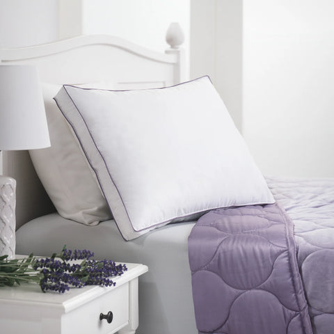 On a white bedside table, is a few lavender flowers. On the bed is a white pristine pillow, with a view of the lavender colored comforter. 