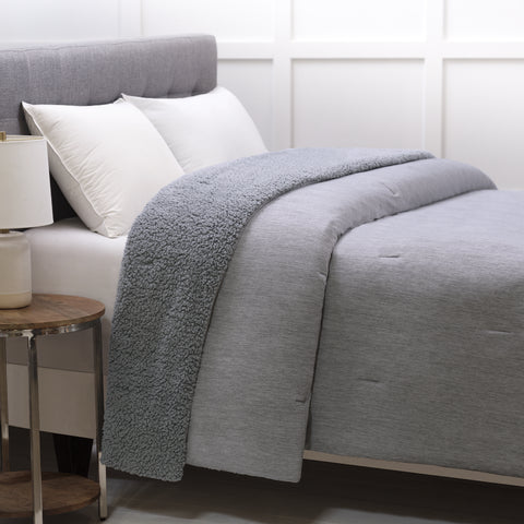 The Reversible Cooling Sherpa Blanket laying on a bed. At the head of the bed is pillows leaning on a head board. This grey bulky blanket looks like a perfectly average blanket but at the blanket is there overlapping on itself and you can see the fuzzy side sticking it's head throw.