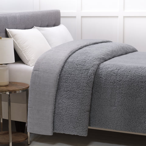 The Reversible Cooling Sherpa Blanket laying on a bed. At the head of the bed is pillows leaning on a head board. Then the blanket is there overlapping on itself. This grey bulky blanket looks almost like one of those fuzzy carpets that one might lay on. Looking extremely comfortable and textured.