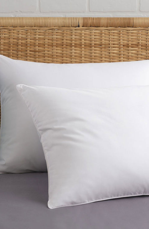 Two PureAssure Allergen Barrier Down Alternative Pillows, one leaning on a light brown whickered headboard, while the other is leaning on that pillow. Both pillows are pristine shade of white.