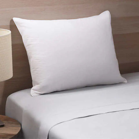 RDS White Duck Down Pillow on a what looks like wood floor as the headboard. It sitting on top of a a bed with white sheets. Just barely in the shot is a wooden bedside table with a lamp that has a black stem and white lamp shade.