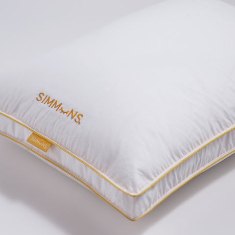 In this image is a close up of corner of the Simmons Gusset Embroidered Pillow on a white background. The Simmons yellow lining around the ends as always. On the side of the pillow is a little yellow tag that says Simmons. On the pillow itself also on the left and the text facing the left is the Simmons' logo. The "o" is an image of a closed eye, looks like a parentheses with four eyelashes.