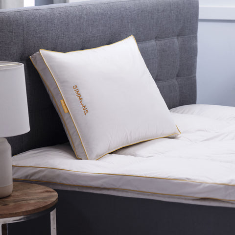In this image is a side view of the Simmons Gusset Embroidered Pillow on a bed. The Simmons yellow lining around the ends as always. On the left side of the pillow is a little yellow tag that says Simmons. On the pillow itself also on the left and the text facing the left is the Simmons' logo. The "o" is an image of a closed eye, looks like a parentheses with four eyelashes.