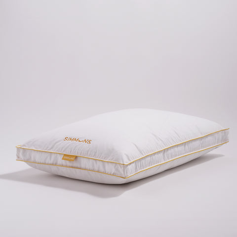 In this image is a side view of the Simmons Gusset Embroidered Pillow on a white background. The Simmons yellow lining around the ends as always. On the side of the pillow is a little yellow tag that says Simmons. On the pillow itself also on the left and the text facing the left is the Simmons' logo. The "o" is an image of a closed eye, looks like a parentheses with four eyelashes.
