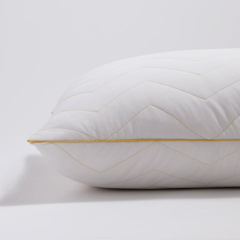 There is a single Simmons Cooling Herringbone Pillow in this image. Getting a side view of the pillow. Laying flat on a white background and zoomed in on the bottom left corner of the pillow. The pillow has the classic Simmons' yellow lining on the edges, with the yellow Simmons' tag on it. The pillow has a zig-zag herringbone quilt pattern and is covered by an "cool to the touch" nylon cover. 