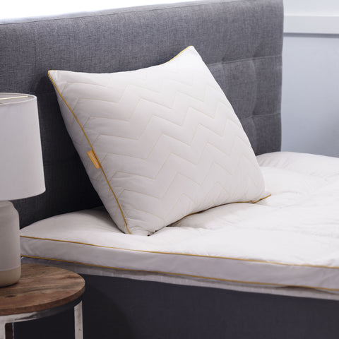There is a single Simmons Cooling Herringbone Pillow in this image. Leaning on the dark gray headboard behind it. The pillow has the classic Simmons' yellow lining on the edges, with the yellow Simmons' tag on it. The pillow has a zig-zag herringbone quilt pattern and is covered by an "cool to the touch" nylon cover. 