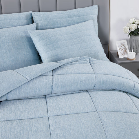In this image, it is a close up of a bed. With a grey headboard. Four light blue pillows leaning on the head board, the pillows on the left are bit out of frame. In front of the pillows, one can see the beginning of a light blue comforter. Just barely in frame on the right of the bed is a grey night stand with a white mug, white picture frame, and white flowers in a zebra printed vase.