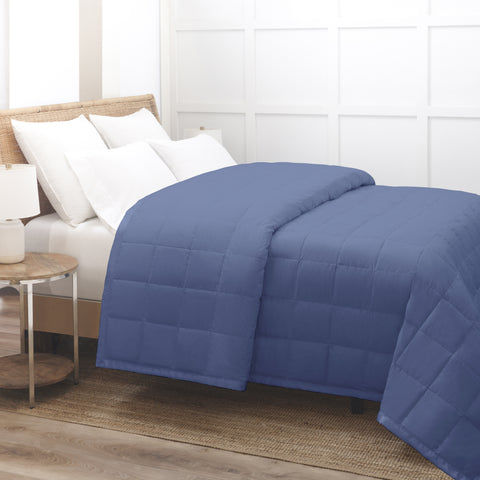 Blue Luxury Down Filled Blanket laying perfectly on top of a bed. We are getting a side view of the bed. The blanket is folded over itself at the head of the bed, yet the blanket is still laying pretty flat. The blanket has squares going all across it.