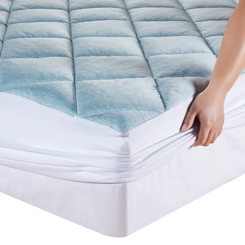 Zoomed into image of a corner of a mattress. With a hand on the right side of the corner putting on a mattress mad. The top of the mattress pad is light bluewith a diamond design running across it.  The side of the mattress pad that wrap around the mattress, are white.