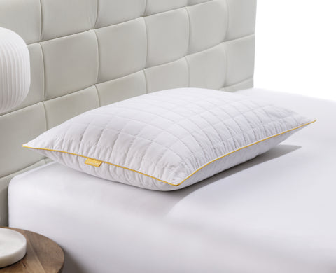 Pillow laying flat on a bed. Getting a side look of the pillow. Squares stitching across the surface. Classic Simmon's Yellow line around the ends and a matching yellow Simmons tag on the left side. 230 thread count quilted cover.