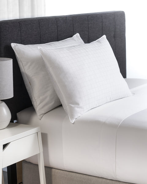 Two 80/20 pillows leaning on a headboard of a bed. With a nightstand beside the bed half out of frame with a lamp on top. The pillows are on twin bed.