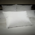 A bed with two pillows leaning on a headboard,. Two more pillows in front of those pillows. Then front and center in front of those four pillows, is a single Perfect White Goose Down Pillow - 50/50 Fill - Medium Plush Support.