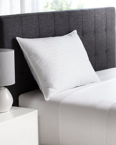 Plush Perfect Gel Fiber Pillow (Medium) (Set of 2), only one of the pillows is pictured here. The whit pillow on a white blanket and white mattress. The single pillow leaning on a a dark grey headboard.