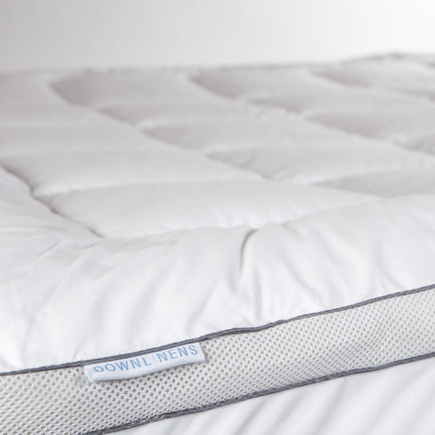 PowerNap Celliant Fiber Blend Mattress Pad it's a view of the side of the mattress pad. We can see on top that we have square stitching and on the side we can see two parallels blue lines with little circles filling the blank space between the lines. A small white tag sticking out with blue text that reads "DOWNLINENS."