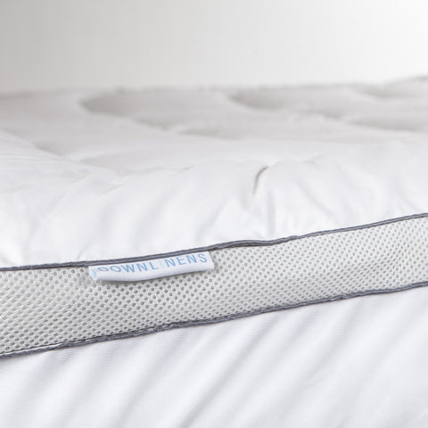 PowerNap Celliant Fiber Blend Mattress Pad it's a view of the side of the mattress pad. We can see on top that we have square stitching and on the side we can see two parallels blue lines with little circles filling the blank space between the lines. A small white tag sticking out with blue text that reads "DOWNLINENS."