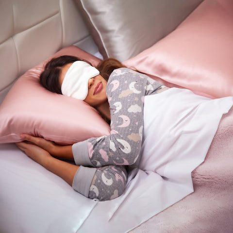 In this image we have a woman in her gray pajamas with moons and clouds on them. She is laying there with the ivory eye mask on, her hands tucked under a pillow with the pink pillow case and her head pressed on that pillow. Beside her is a pillow in silver pillow case leaning on the white headboard, with a pillow laying flat in front of it in a pink pillow case.