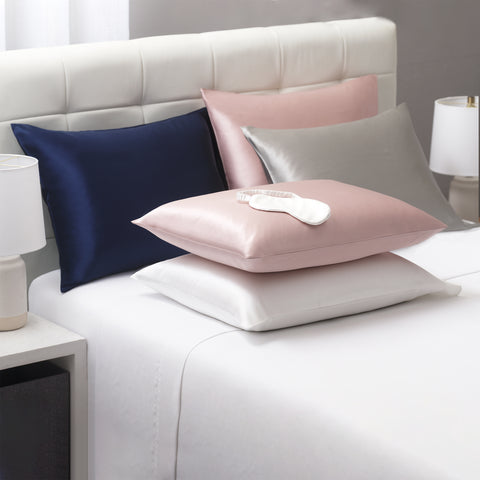 On a white bed, we have all sorts of Silk Pillow Cases. On the left against the headboard is a navy blue silk pillow case on a a pillow. Beside it is a pink one, with a silver on leaning on top of the pink. In front of the navy one is white one laying flat with another pink on tip. There is an ivory silk mask laying on top of the pink one.