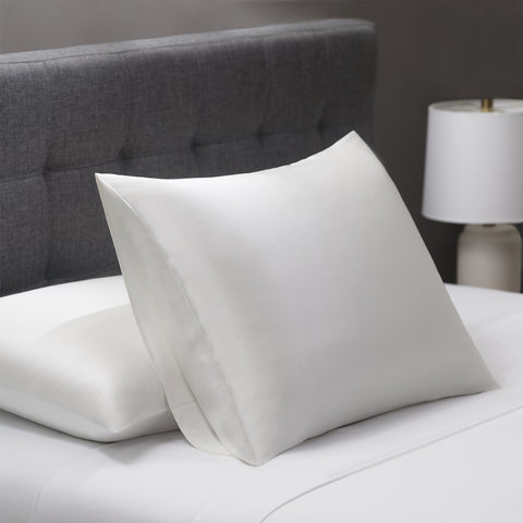 On a white bed with a dark grey head board, lay a couple pillows. Both pillows are inside the ivory silky pillow case. One pillow lays flat on the white bed, close to the headboard. The other one leans on the flat pillow propping itself up.