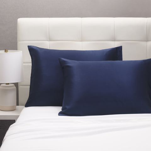 On a white bed is a few pillows in a navy silk pillows case. all leaning on a white headboard. The silky pillow cases seem as though they shimmer. 