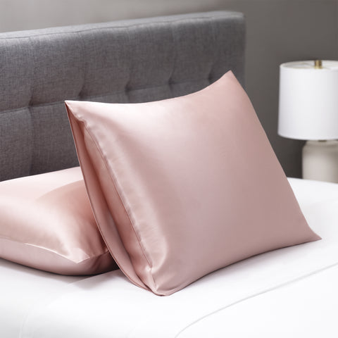 On a white bed with a dark grey head board, lay a couple pillows. Both pillows are inside the pink silky pillow case. One pillow lays flat on the white bed, close to the headboard. The other one leans on the flat pillow propping itself up.