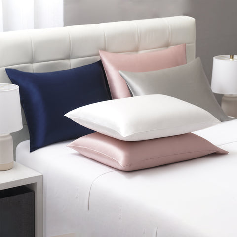 On a white bed, we have all sorts of Silk Pillow Cases. On the left against the headboard is a navy blue silk pillow case on a a pillow. Beside it is a pink one, with a silver on leaning on top of the pink. In front of the navy one, is  another pink on laying flat with a white one on top.